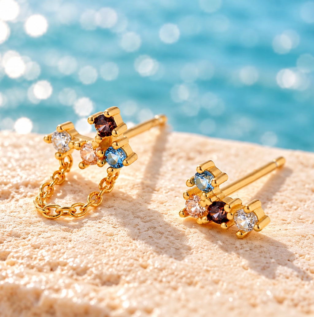 Asymmetric multicolour gold stud earrings with one delicate loop chain by a turquoise lake