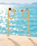 Asymmetric gold chain drop earrings with colourful stones against a turquoise lake