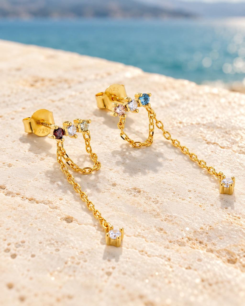 Colourful gold chain earrings lying on pale stone by blue water