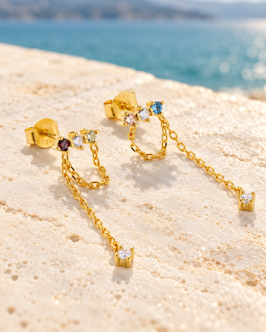 Colourful gold chain earrings lying on pale stone by blue water